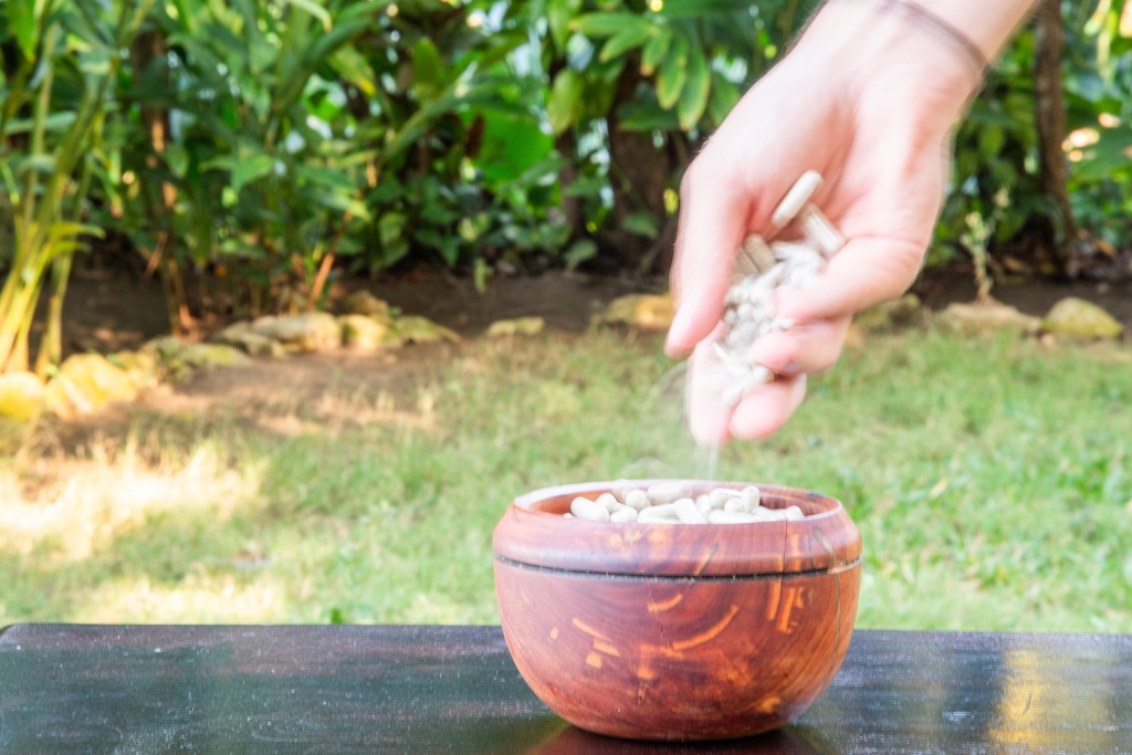 Hand dropping psilocybin mushroom capsules into a wooden bowl on table with green plants and grass in the background.