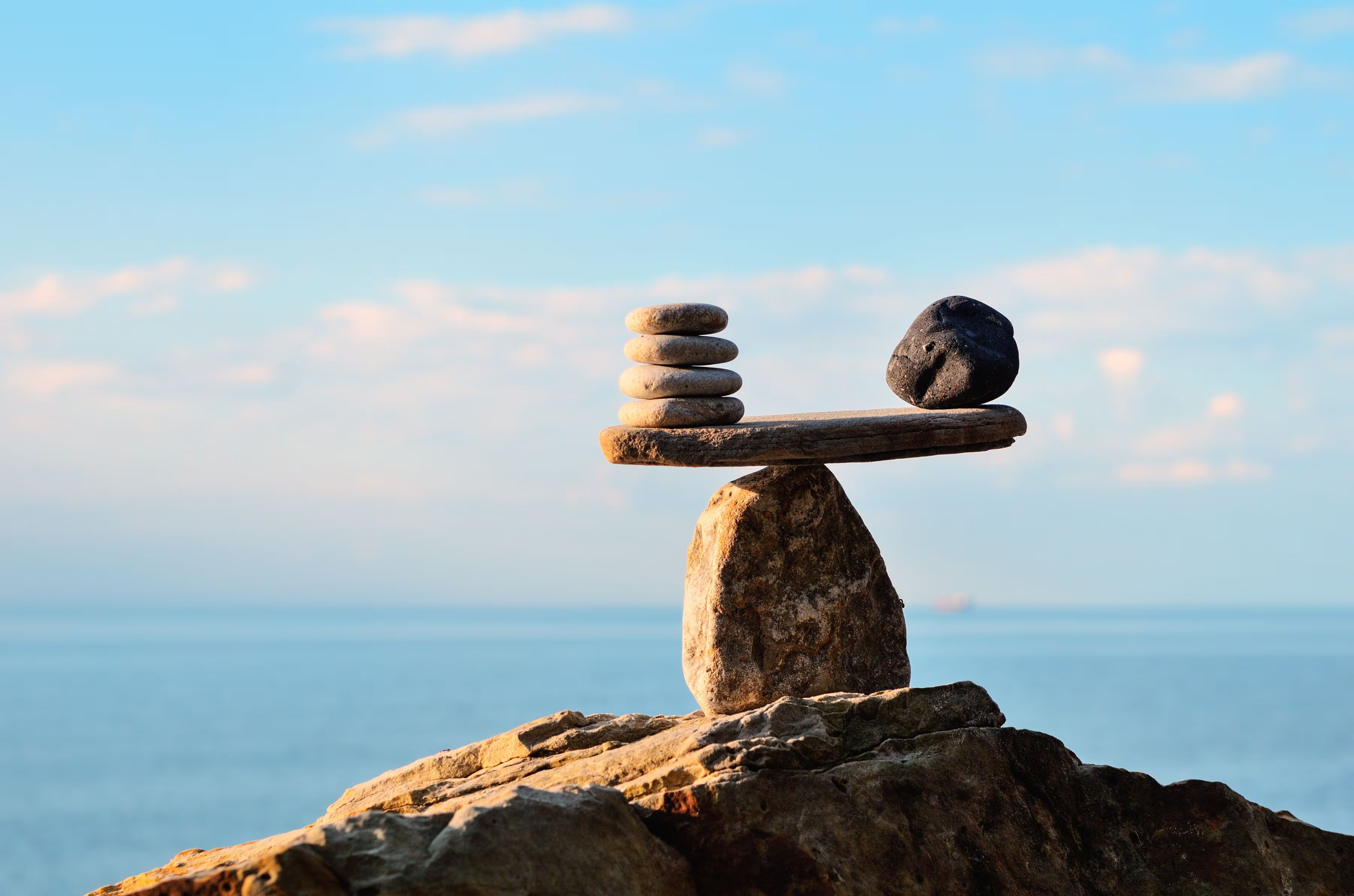 Seven stones in perfect balance on top of a larger rock with clear sky and sea in the background.