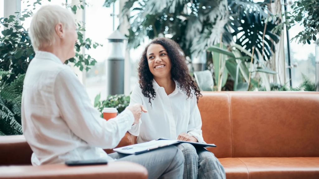 Two women working and laughing while sitting on leather couches with green plants in background.