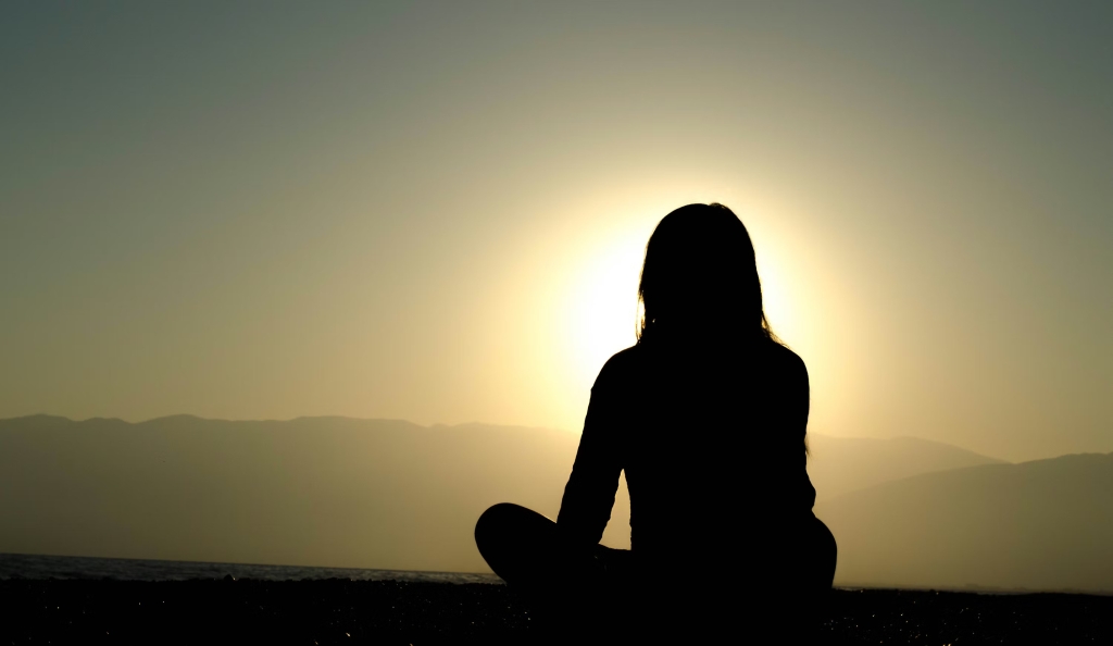 A silhouette of a woman sitting with her legs crossed in meditation as the sun goes down.