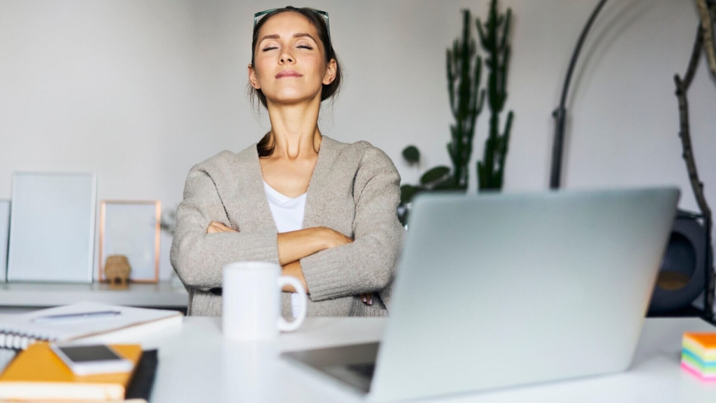 Woman at work taking a mindfulness break at her desk, sitting peacefully with eyes closed.