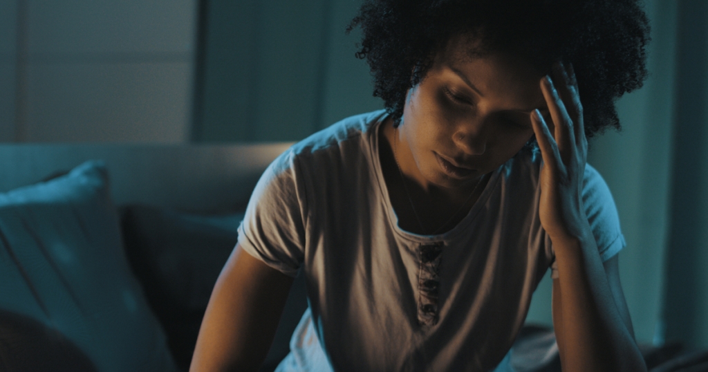 A woman sitting on the edge of her bed in low ligh unable to sleep.